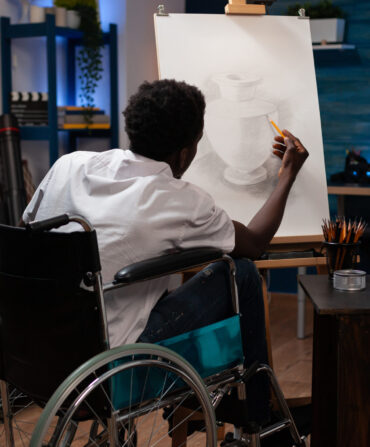 Young african american male artist user of wheelchair having good time making pencil drawing on canvas easel of pot. Man with artistic skills creating artwork at home art studio.